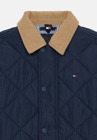 Quiltet marineblå jakke med en beige kordfløyelskrage, knappeåpninger og en liten brodert logo på brystet. Stripete indre fôrdetalj.