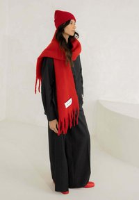 Rode gebreide muts en oversized rode sjaal van zachte wol, gedragen met een zwart gestreept pak en rode schoenen. De sjaal heeft franjes als accent.