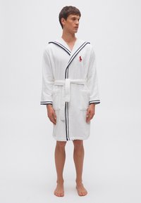 Άνδρας όρθιος, ξυπόλυτος, φοράει λευκή ρόμπα με ναυτικό μπλε επένδυση και κόκκινο κεντητό λογότυπο σε απλό ανοιχτό φόντο.