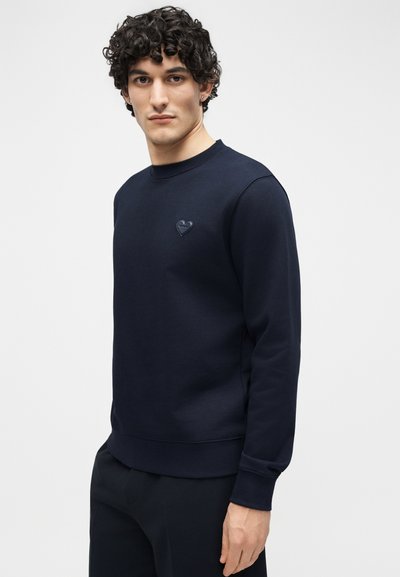 Νέος άνδρας με σγουρά μαλλιά, που φοράει μπλε σκούρο φούτερ με ένα μικρό έμβλημα καρδιάς στο στήθος, στέκεται μπροστά σε ένα απλό φόντο.