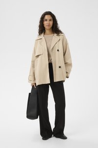 Bézs színű, bő szabású ballonkabát két nagy fekete gombbal, világos pulóverrel és fekete, széles szárú nadrággal párosítva. Egy fekete válltáskát tart a kezében.