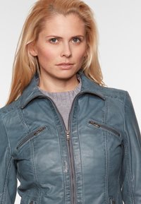 Teal leren jack met een ritssluiting aan de voorkant, een staande kraag en twee ritszakken op de borst. Beschikt over textuurpanelen en stikdetails.