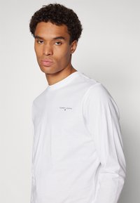 Camisa de manga longa branca feita de algodão com decote redondo, apresentando um pequeno logo preto "TOMMY JEANS" na área do lado esquerdo do peito.