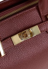 Close-up van een kastanjebruine, gestructureerde leren handtas met een gouden metalen sluiting en ritsdetails.