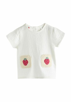 Chemise blanche à manches courtes en tissu texturé, ornée de deux patchs brodés de fraises sur fond beige carré.