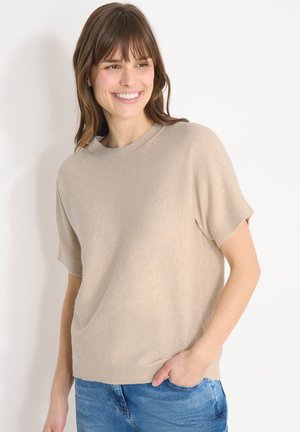 Femme souriante aux cheveux bruns mi-longs portant un pull beige à manches courtes et un jean bleu, debout devant un fond blanc uni.