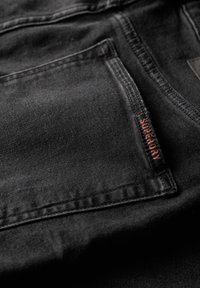 Tejido de mezclilla negra con un bolsillo frontal que lleva una etiqueta roja de "Superdry", detalles de costura sutiles y una textura suave.