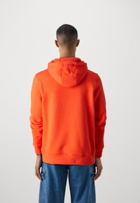 Persona di spalle che indossa un felpa arancione brillante e pantaloni cargo in denim blu su uno sfondo chiaro e semplice.