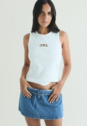 Femeie cu păr lung și închis la culoare, purtând un top alb fără mâneci și o fustă mini din denim albastru, ajustând talia fustei cu ambele mâini.