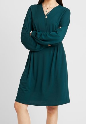 Turquoise wikkeljurk met lange mouwen, gemaakt van gladde stof. Heeft een V-hals en een losse pasvorm, eindigt boven de knieën.