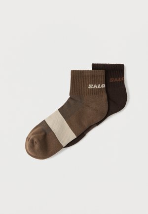 Paar knöchellanger Socken, eine dunkelbraun und eine braun mit einem beigen Streifen, beide mit gerippten Bündchen und dem Text "SALO" nahe der Oberkante.