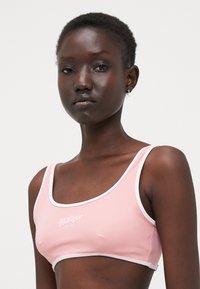 Lys rosa cropped bh-top med hvit kant, med en logo trykket i midten. Glatt tekstur og firkantet utringning.