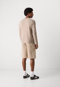 Beiger Strickpullover, lange Ärmel, runder Ausschnitt, kombiniert mit beigen Shorts, schwarzen Sneakers mit weißen Streifen und weißen Crew-Socken.