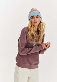 Fioletowa bluza z okrągłym dekoltem z wyszywanym logo "The Nomad", noszona z beżowo-niebieską dzianą czapką. Miękki materiał, luźny krój.
