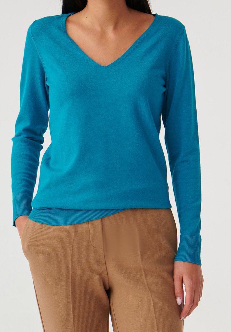 Femme portant un pull à col en V manches longues bleu sarcelle rentré dans un pantalon camel taille haute, les mains dans les poches.