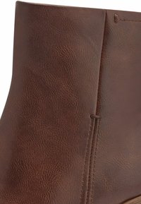 Bottines en cuir marron avec une finition texturée, des coutures verticales, et un contour lisse. La semelle est en bois de couleur claire.