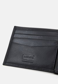 Porte-cartes en cuir noir avec plusieurs fentes et logo en relief. Surface texturée avec des détails de couture visibles. Design compact et rectangulaire.