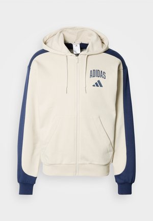 Kapuzensweatshirt zum Reißverschluss in Creme mit marineblauen Ärmeln. Verfügt über Fronttaschen und das "Adidas"-Logo auf der Brust gedruckt. Weiche, Fleece-Textur.