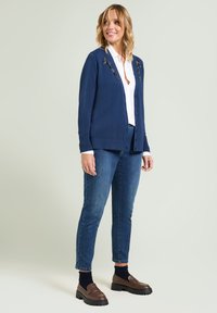 Marineblauwe cardigan met knopen aan de voorkant, geribde manchetten en versierde details op de schouders, gecombineerd met een blauwe spijkerbroek en bruine loafers.