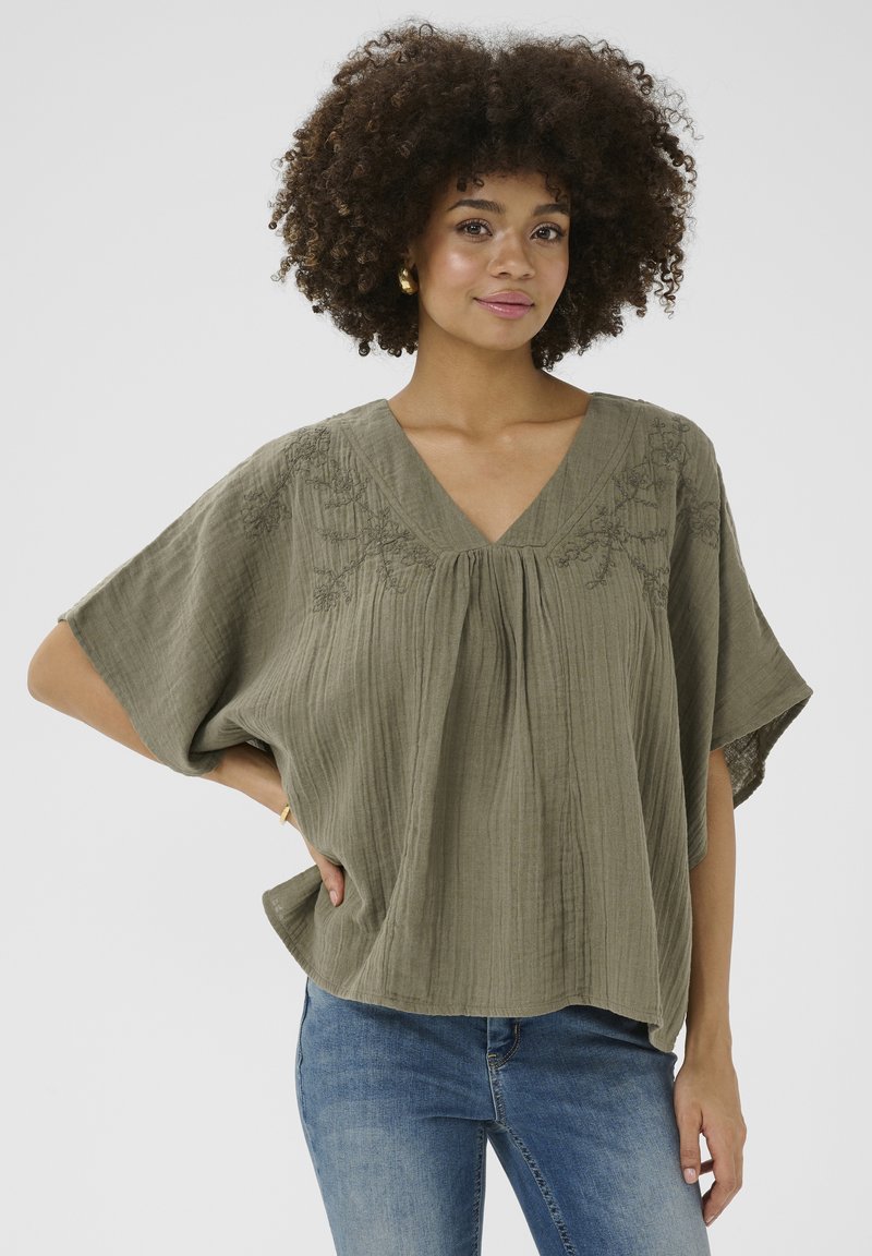 Olíva zöld blúz rövid ujjakkal, V-nyakkivágással és texturált anyagból. Díszítő hímzés a vállakon, részletgazdag megjelenést kölcsönöz. Farmerrel viselve.
