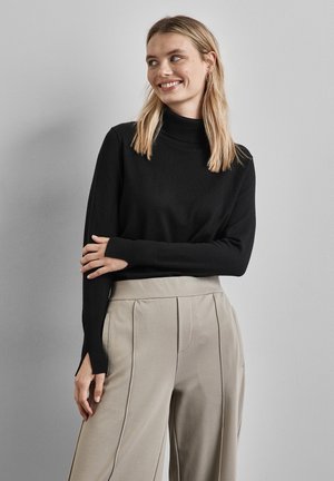 Maglione nero a collo alto in tessuto leggero con maniche lunghe, abbinato a pantaloni beige a vita alta plissettati per un look elegante.