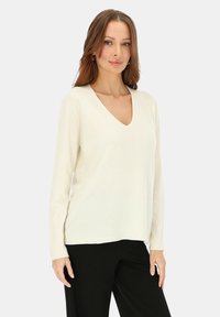 Kremowy sweter z dekoltem w serek, z długimi rękawami, miękką fakturą i luźnym krojem, pokazany na czarnych spodniach. Prosty, minimalistyczny design.