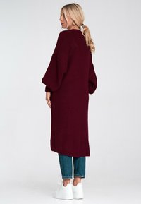 Bordowy, dziany długi sweter z szerokimi rękawami, prążkowaną teksturą i bocznymi rozcięciami, w połączeniu z dżinsami do kostek i białymi sneakersami.