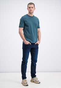 Žalsvai mėlynas marškinėlių modelis su tamsiai mėlynomis džinsomis. Modelis dėvi smėlio spalvos sportinius batelius. Audinys atrodo minkštas ir lygus.