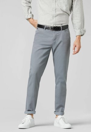 Man draagt lichtgrijze broek, zwarte riem, gestreept overhemd in de broek gestopt en witte sneakers, staand met één hand in de zak.
