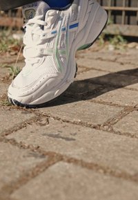 Balti sportiniai batai su tinkleliu, žaliais ir mėlynais akcentais, bei guminėmis padangomis, padėti ant akmeninio grindinio.