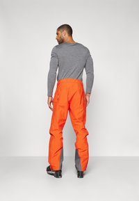 Orangene Schnee-Hosen mit grauen Akzenten, Cargo-Taschen an den Seiten und einer schmalen Passform an den Knöcheln. Tragen mit einem grauen Langarmshirt.