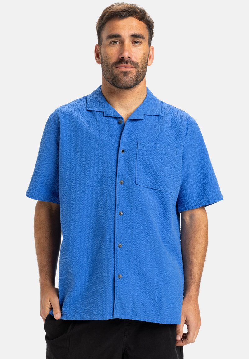 Camisa de manga corta, con botones, en tejido texturizado azul brillante, con un bolsillo en el pecho y cuello con muesca. Presenta botones oscuros.