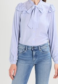 Hellblau gestreifte Bluse mit Rüschenschultern und einer Krawattenkragen, kombiniert mit hellblauen Jeans, die einen lässigen, maßgeschneiderten Look zeigen.