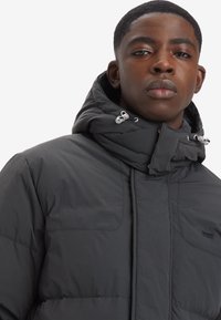 Czarna kurtka puchowa z wysokim kołnierzem i sznurkiem do regulacji. Posiada pikowaną fakturę, dwie kieszenie na piersi oraz subtelne logo po lewej stronie.