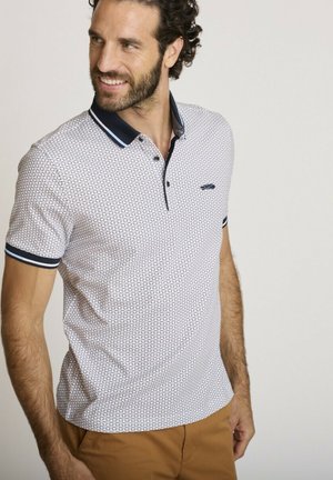 Homme aux cheveux bouclés et à la barbe, portant un polo blanc à motifs avec un col bleu marine et un pantalon beige, souriant et regardant vers la gauche.