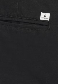 Gros plan sur un tissu noir montrant une poche fendue horizontale avec une étiquette blanche rectangulaire de la marque Jack & Jones cousue au-dessus.