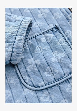 Jachetă din denim matlasat în albastru deschis cu un model floral subtil. Are un buzunar cu margini rotunjite și material texturat.