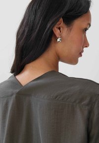 Haut vert olive foncé avec un décolleté en V et une coupe décontractée, présentant une texture lisse et un détail de couture sur les épaules. Boucles d'oreilles en argent visibles.