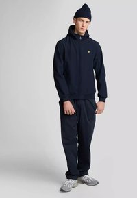 Felpa con zip di color blu navy con cappuccio annesso e polsini a costine, abbinata a pantaloni coordinati. Presenta un logo giallo sul petto. Indossata con sneakers grigie chiare.