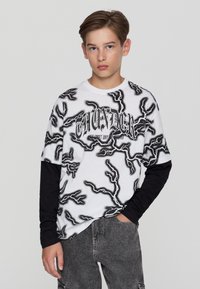 Nastolatek z krótkimi włosami, ubrany w biało-czarną koszulkę z wzorem błyskawic na czarnych długich rękawach i szarych dżinsach, stojący z jedną ręką w kieszeni.