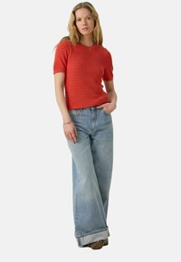 Haut rouge à manches courtes en tricot avec un design texturé, associé à un jean évasé bleu clair, offrant une coupe décontractée et des manches retroussées.