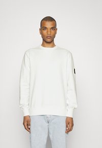 Tricou alb cu guler rotund, manșete și tiv ribbate. Prezintă o patch pe mâneca stângă. Purtați-l cu blugi albaștri deschiși.