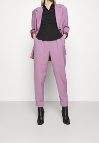 Lila op maat gemaakte blazer met bijpassende broek, gecombineerd met een zwart button-up shirt. Gestructureerde stof, licht oversized pasvorm, gestyled met zwarte enkellaarsjes.