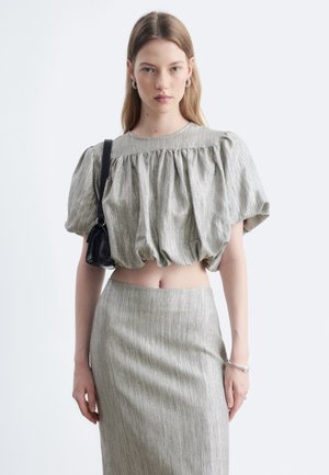 Jeune femme aux cheveux lisses portant un crop top brillant à manches bouffantes et une jupe taille haute assortie, tenant un petit sac à bandoulière noir.