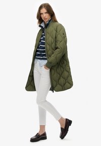 Cappotto trapuntato verde oliva con polsini a coste, abbinato a una maglietta a righe blu navy e pantaloni bianchi, completato da mocassini borgogna.