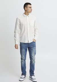 Camisa blanca de botones en tejido ligero, con bolsillo frontal, combinada con jeans azules desgastados y zapatillas negras y blancas a rayas.