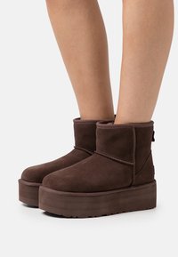 Brown suede ankle boots with a round toe, layered platform sole, and stitched detailing. Features a low cuff and back loop for easy wear.