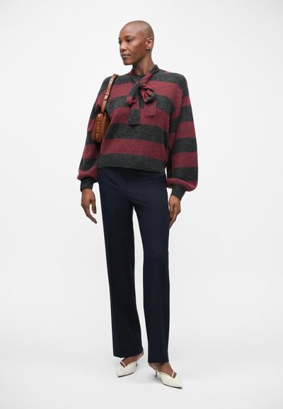 Pull rayé rouge et gris avec un détail nœud papillon, associé à un pantalon bleu marine et des talons blancs pointus. Petit sac à main marron sur l'épaule.