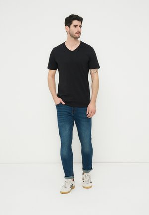 Άνδρας με κοντά σκούρα μαλλιά, φορώντας μαύρο T-shirt με V λαιμόκοψη, μπλε τζιν, λευκά αθλητικά παπούτσια, στέκεται μπροστά σε ένα απλό ανοιχτό φόντο.