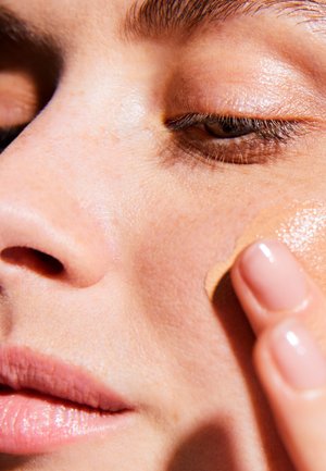 Eine Nahaufnahme eines Gesichts, das glatte Haut zeigt, auf der eine leichte Foundation aufgetragen wird. Subtile Texturen und ein natürlicher Hautton sind deutlich sichtbar.